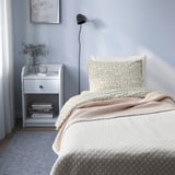 Dormitorio acolledor con cama individual e cuberta de cama PRAKTVIAL, mesa de noite con libros e flores, lámpada de parede.