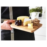 Unha persoa que sostén unha táboa de corte de madeira con queixo, uvas e crackers. O taboleiro ten un mango para facilitar o transporte.