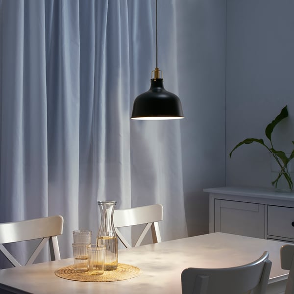 A luz colgante negra colga sobre a mesa de comedor branca, contrastando o deseño industrial con cadeiras modernas e cortinas de luz.