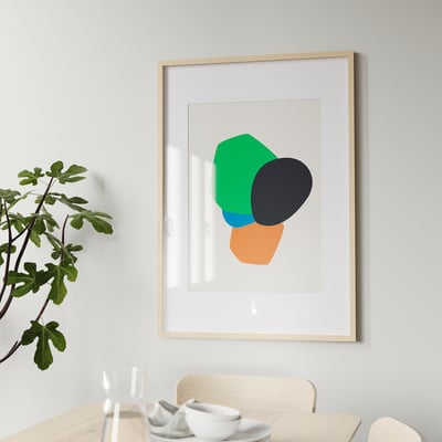 A arte abstracta enmarcada colga sobre unha mesa de comedor de madeira con cadeiras e pratos brancos. Unha planta está situada á esquerda.