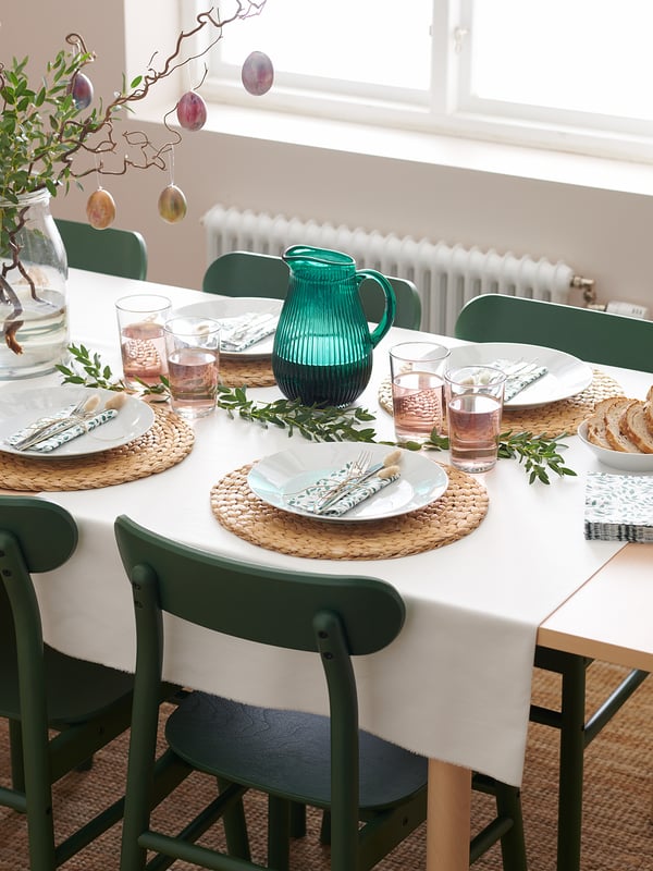 Mesa de comedor conxunto con pratos brancos, cadeiras verdes, xerra de vidro, servilletas e pratería. Lugares sinxelos e elegantes.