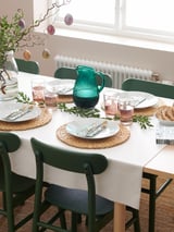 Mesa de comedor conxunto con pratos brancos, cadeiras verdes, xerra de vidro, servilletas e pratería. Lugares sinxelos e elegantes.