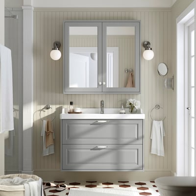 Baño moderno con vaidade gris RUTSJÖN, espello, ducha e lavabo de cerámica. Acentos brancos, luces de parede e aneis de toalla cromada.