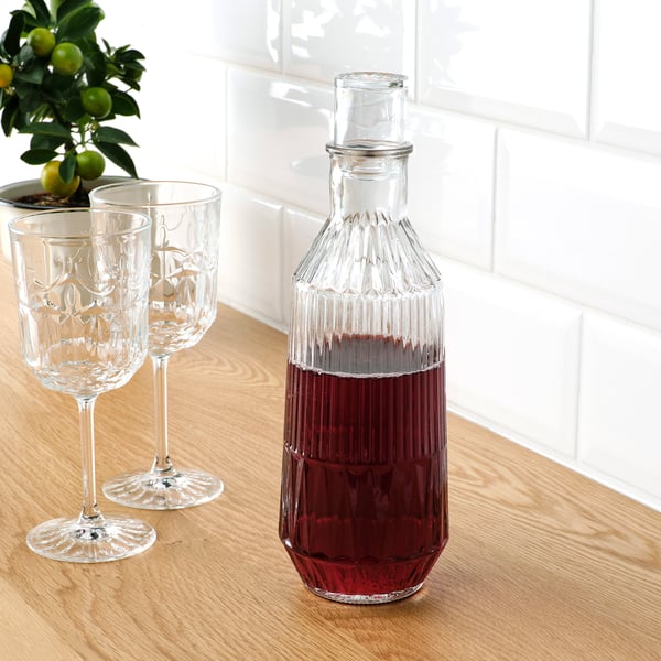 Caraffa de vidro transparente con bebida vermella, dous vasos correspondentes, planta de cítricos sobre mesa de madeira contra parede de azulexos brancos.