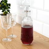 Caraffa de vidro transparente con bebida vermella, dous vasos correspondentes, planta de cítricos sobre mesa de madeira contra parede de azulexos brancos.