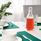 Unha mesa de comedor con cuncas brancas, servilletas verdes e unha carafa de líquido vermello.