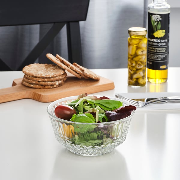 A cunca de vidro transparente chea de ensalada séntase nunha mesa branca, acompañada de crackers e condimentos.