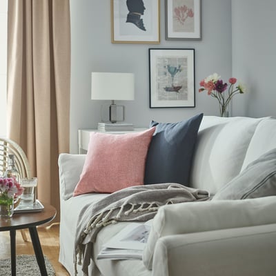 Sala de estar luminosa con sofá branco, almofadas vermellas e azuis, mesa lateral de madeira, lámpada, flores e cortinas grises.