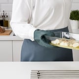 A persoa que leva unha blusa branca e un mandil gris usa un guante de forno de silicona verde para manexar un prato de forno desde o forno, garantindo a protección e o agarre das mans.