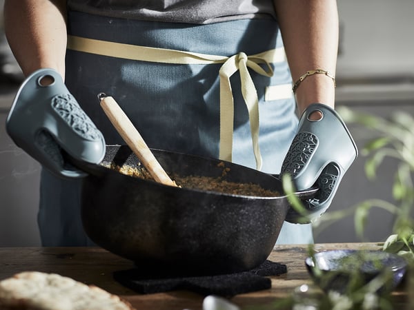 Persoa en mandil e guantes de forno mantén tixola con comida, cociñando.