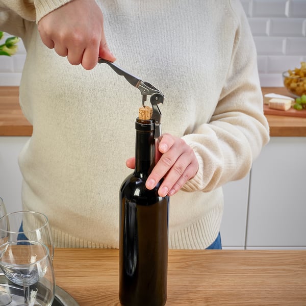 A persoa abre a botella de viño usando un parafuso de corcho sobre a mesa de madeira. Dúas copas de viño nas proximidades.
