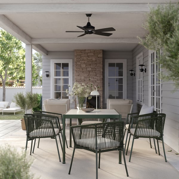 Patio exterior con cadeiras verdes SEGERÖN, mesa rectangular e vaso branco. Ventilador de teito, casa branca e muro de ladrillo.