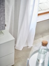 As cortinas brancas colgan xunto a un armario branco. A luz filtra suavemente a través da textura de liño de tecido 100% algodón, creando sombras suaves.