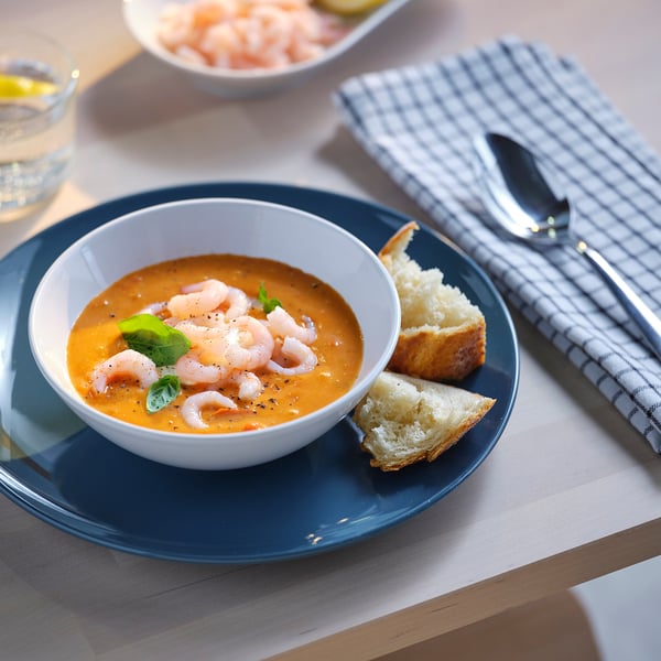 Unha cunca de sopa de camaróns de laranxa con pan, colocada nun prato azul con pratería nunha servilleta a cadros.
