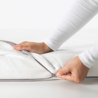 Unha persoa pon un colchón de berce branco, destacando a súa cuberta fácil de retirar e lavable na máquina para limpeza e seguridade.