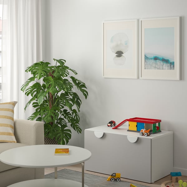 Pequeno banco de almacenamento para nenos gris con xoguetes preto dunha planta verde, arte enmarcada na parede.