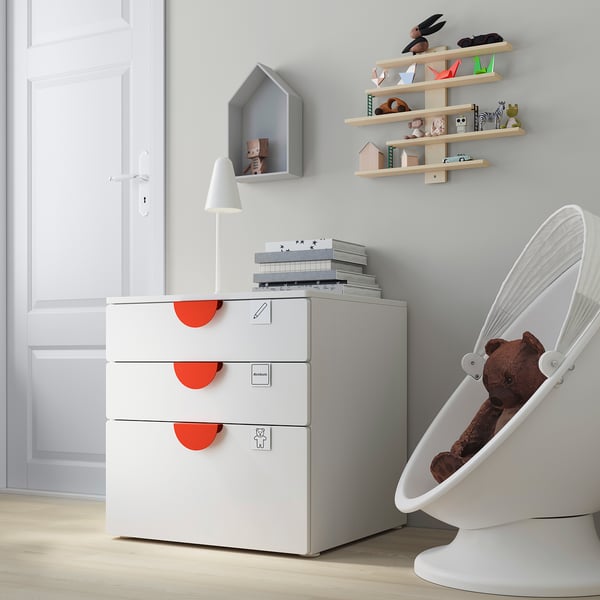 Habitación moderna para nenos cun cofre branco SMÅSTAD, estantes de madeira clara e unha cadeira colgante branca cun oso de peluche.