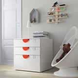 Habitación moderna para nenos cun cofre branco SMÅSTAD, estantes de madeira clara e unha cadeira colgante branca cun oso de peluche.