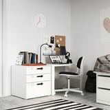 Pequeno escritorio branco no dormitorio, cadeira gris, portátil, libros, lámpada, mochila, alfombra a raias, reloxo, carteis.
