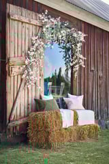 Entrada rústica decorada cun arco floral branco. Balas de feno con almofadas verdes e rosas sentan diante.