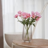 Un vaso claro e nervado con flores artificiais rosas colocado sobre unha mesa de madeira xunto a unha fiestra. As flores traen cores vibrantes e decoración ao espazo.