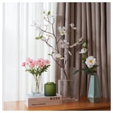 Tres vasos con flores nunha mesa de madeira preto dunha cortina; unha ten flores rosas, outra ten flores brancas e unha vela verde sentada entre elas.