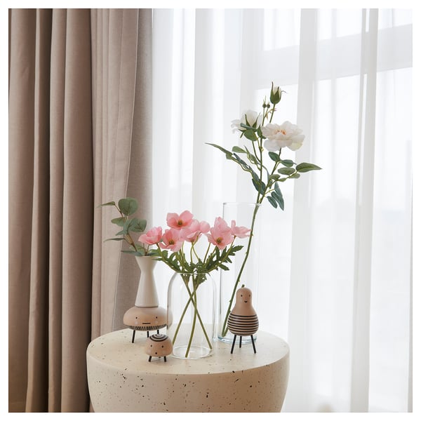 Tres vasos con flores sobre unha mesa, dous de vidro claro, un de cerámica branca. Cortinas en fondo.