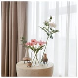 Tres vasos con flores sobre unha mesa, dous de vidro claro, un de cerámica branca. Cortinas en fondo.