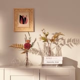 Un bouquet elegante nun vaso transparente senta nun armario branco, engadindo cor e vida. Por riba del, colga un cadro de marco tecido.