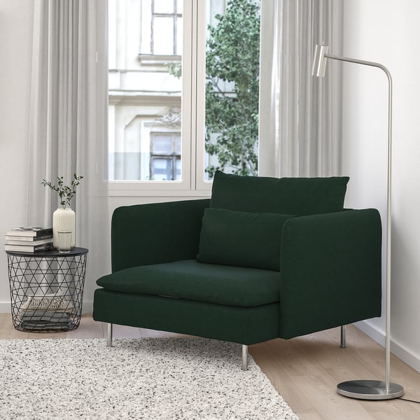 Acolledora cadeira verde SÖDERHAMN por fiestra. Asentos modernos e profundos, patas metálicas. Mesa final ordenada con libros e un vaso. Alfombra suave debaixo. Lámpada de chan á dereita.
