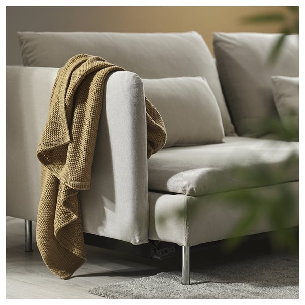 Sofá-cama beige moderno con patas metálicas, grandes coxíns e unha manta acolledora sobre o repousabrazos.