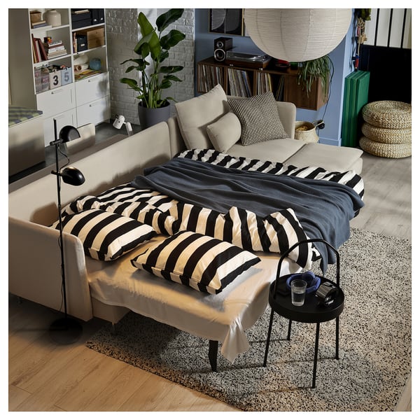 Sala de estar moderna con sofá-cama beige, almofadas de raias brancas e negras, mesa lateral, alfombra, plantas e estantes con libros e decoración.