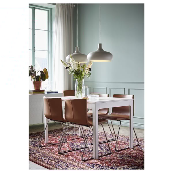 Comedor moderno con mesa branca, cadeiras marróns e luces colgantes. Paredes azuis, alfombra floral, flores sobre a mesa.