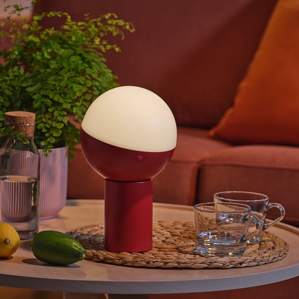 Lámpada de mesa moderna con sombra de medio globo. Espazo acolledor cunha base vermella. Rodeada de cuncas e froitas nunha mesa lateral.