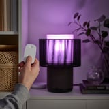 Unha man sostén un remoto, controlando unha lámpada elegante cun paralume cilíndrico, emitindo luz púrpura.