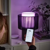 O teléfono controla a lámpada púrpura a través da aplicación. A lámpada ten sombra raiada, senta nun estante púrpura con plantas e libros.