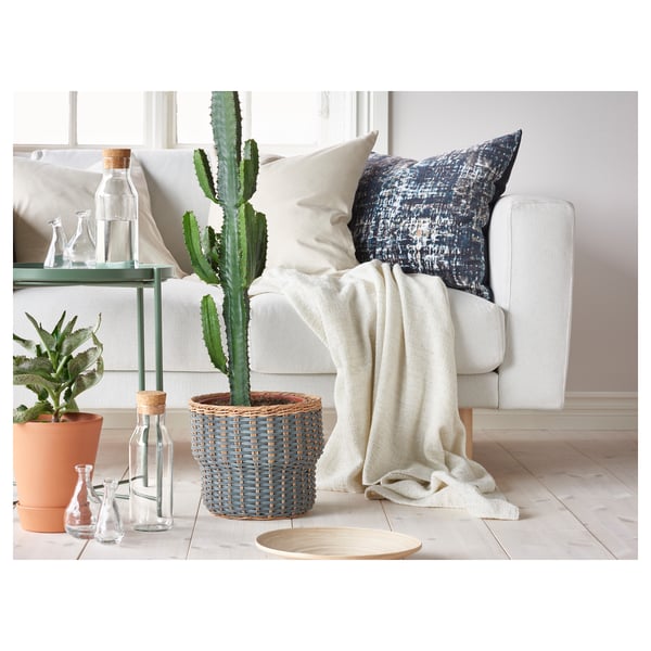 Sala de estar moderna con sofá branco, almofada con patróns azuis, vasos de vidro ben dispostos, unha cesta de vimbio con cactus e unha planta de esquina.