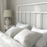 Unha cama ben feita cun marco metálico branco, cabeceira curva e múltiples almofadas e mantas brancas, suxerindo confort e soporte.