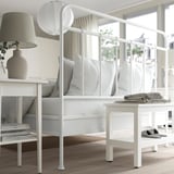 Cama branca moderna con cabeceira curva, roupa ordenada, mesa de noite e lámpada, amplo almacenamento.