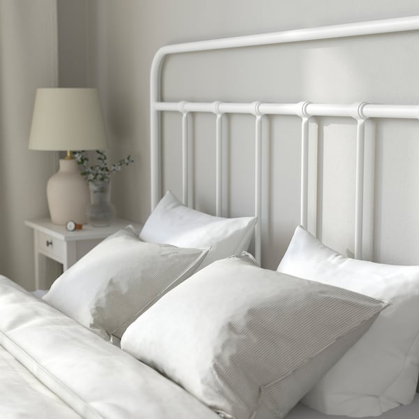 Cama de metal branco con cabeceira curva, roupa branca, almofadas de raias e mesa de noite con lámpada, reloxo e vaso.