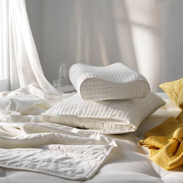 Dúas almofadas brancas apiladas nunha cama con roupa branca e amarela, preto dunha xanela con cortinas transparentes.