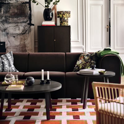 Sala de estar con sofá de veludo marrón, manta verde, mesas de café, velas, libros, alfombra xeométrica, cadeira e armario.