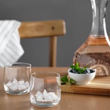 Dous vasos cheos de cubos de xeo sentados nunha mesa de madeira. Xunto a eles, unha carafa de líquido de cor rosa e unha cunca de arandos mesturados e menta.