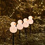Catro decoracións en forma de home de neve iluminanse, proxectando un brillo cálido nunha cama de xardín, creando un ambiente festivo pola noite.
