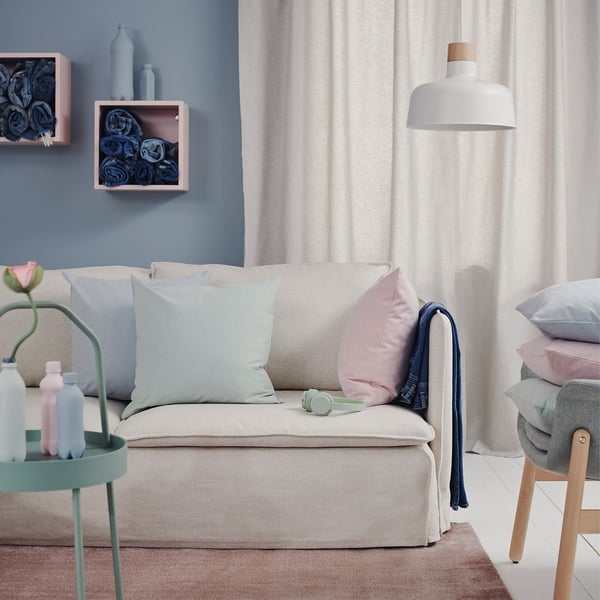 Sala de estar con sofá beige, coxíns azuis claros e rosas, lámpada branca colgada, parede azul, cortina e mesa lateral verde con botellas.