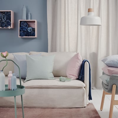 Sala de estar con sofá beige, coxíns azuis claros e rosas, lámpada branca colgada, parede azul, cortina e mesa lateral verde con botellas.