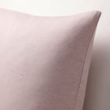 Primeiro plano de cuberta de coxín rosa claro suave e texturizada contra unha superficie correspondente. Detalle de costuras e tecidos sutil efecto slub visible.