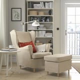 Recuncho acolledor: cadeira de brazos beige, ottoman, mesa lateral con cunca e revistas. Gran estante detrás, iluminado por unha lámpada de pé. Porta con vista á fiestra.