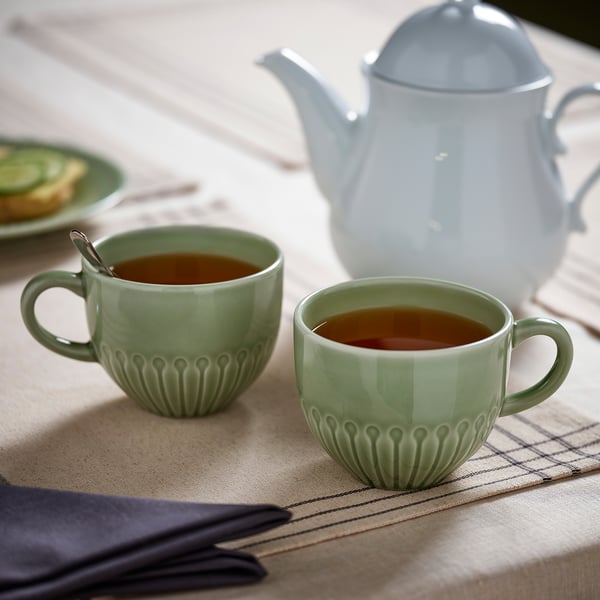 Dúas cuncas de té verde menta con mangos están nun pano de mesa beige xunto a unha teteira branca. As cuncas están cheas de té.