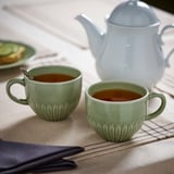 Dúas cuncas de té verde menta con mangos están nun pano de mesa beige xunto a unha teteira branca. As cuncas están cheas de té.